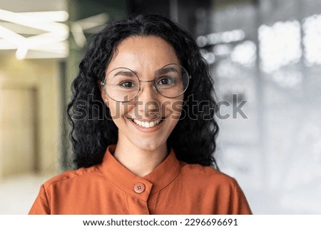 Close up photo portrait of beautiful Latin American woman with curly hair and glasses, businesswoman inside office building smiling and looking at camera