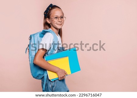 Happy smiling smart little teenager blond kid school girl of 10 years old in casual clothes with backpack holding spiral notebook isolated over beige background studio portrait. Back to school.