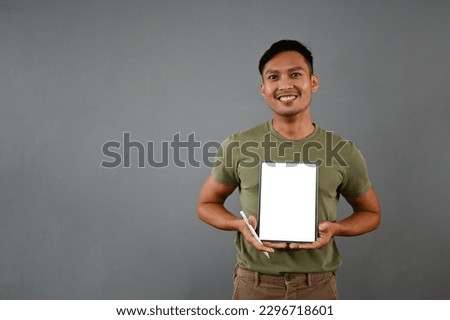 Handsome millennial Asian man holding a digital tablet, showing a tablet white screen mockup while standing on an isolated gray background.