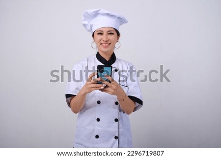 Photo of happy and smiling female Asian chef holding and using mobile phone on isolated white background