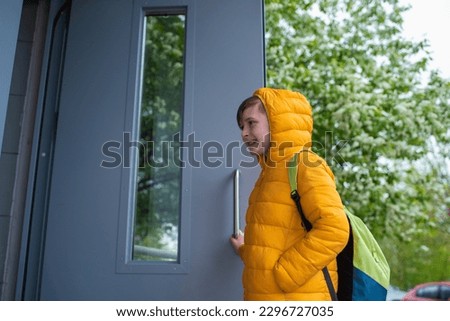 a boy in a jacket with a hood on his head, with a backpack, opens the door to the entrance of a residential building in the spring