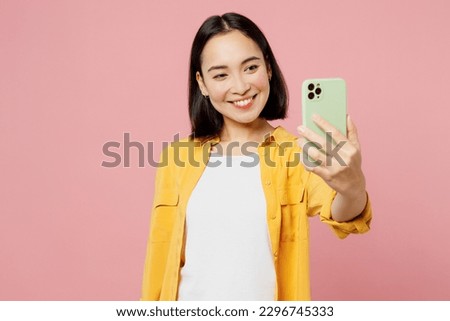 Young happy woman of Asian ethnicity wear yellow shirt white t-shirt doing selfie shot on mobile cell phone post photo on social network isolated on plain pastel light pink background studio portrait