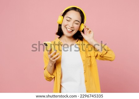 Young fun woman of Asian ethnicity wear yellow shirt white t-shirt headphones listen to music use mobile cell phone isolated on plain pastel light pink background studio portrait. Lifestyle concept