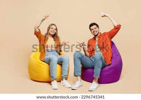 Full body young couple two friends family man woman wear casual clothes together sit in bag chair point index finger between them on area isolated on pastel plain light beige color background studio Royalty-Free Stock Photo #2296745341