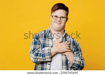 Young smiling thankful man with down syndrome wearing glasses casual clothes look camera put folded hands on heart isolated on pastel plain yellow color background. Genetic disease world day concept Royalty-Free Stock Photo #2296745439