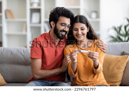 Portrait Of Happy Young Indian Couple Holding Positive Pregnancy Test While Sitting On Couch At Home Together, Eastern Husband Embracing His Joyful Wife, Celebrating Awaiting Baby, Copy Space Royalty-Free Stock Photo #2296761797