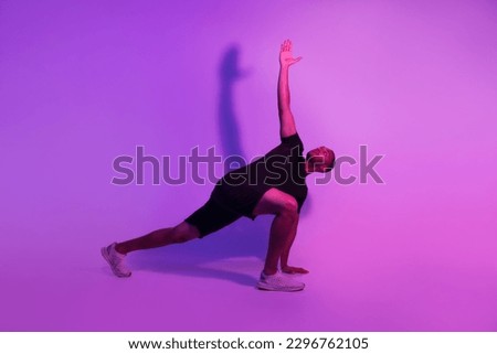 Black Fitness Man Practicing Yoga Standing In Revolved Side Angle Pose Over Purple Neon Studio Background. Sporty Guy Exercising And Stretching Wearing Fitwear. Full Length Shot