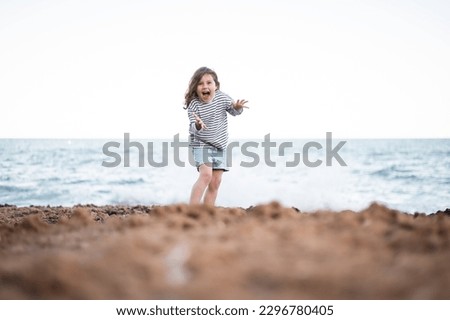 Full body ground level of funny little girl in striped sweatshirt and shorts playing on beach near splashing water from waves against horizon line