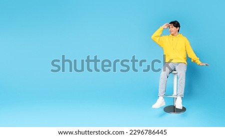Look There. Asian Teen Boy Looking Aside Holding Hand At Forehead Posing Sitting In Chair Over Blue Studio Background. Guy Looks At Empty Space For Text. Offer Advertisement. Full Length, Panorama