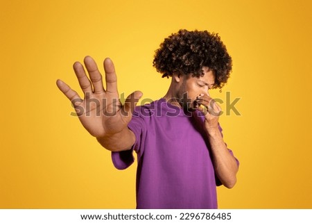 Unhappy adult african american curly man in purple t-shirt covering his nose, making stop sign with hand isolated on yellow studio background. Disgust emotion, stink, bad smell, ad and offer