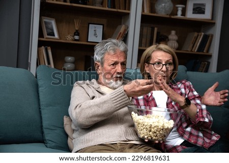 Mature married couple entertaining observing terrified TV program. Senior husband and wife watching horror movie or film at home eating popcorn and making scary facial expressions