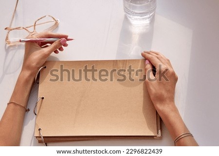 Woman is writing by her left hand on a craft paper album. Glasess and glass of water with hard shadows. Album mockup. Conceptual photo of international left handers day. Copy space. View from above. Royalty-Free Stock Photo #2296822439