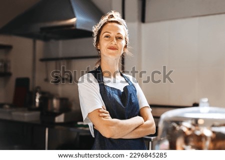 Female cafe owner, showing confidence in her small business, stands in front of the camera with arms crossed. Woman wearing an apron, proudly displays her role as a hospitality entrepreneur. Royalty-Free Stock Photo #2296845285