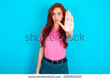 young redhead woman wearing pink T-shirt over blue background shows stop sign prohibition symbol keeps palm forward to camera with strict expression