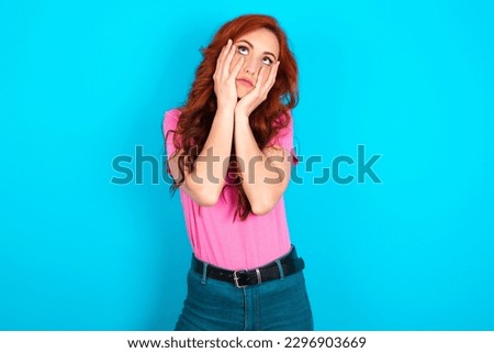 young redhead woman wearing pink T-shirt over blue background keeps hands on cheeks has bored displeased expression. Stressed hopeless model