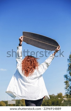 A girl in a white shirt holds a skateboard over her head against a blue sky. Female figure from the back with a surf skate in summer