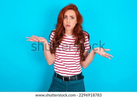 Frustrated young redhead woman wearing striped T-shirt over blue background feels puzzled and hesitant, shrugs shoulders in bewilderment, keeps mouth widely opened, doesn't know what to do. Royalty-Free Stock Photo #2296918783