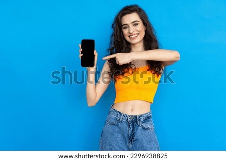 Portrait of an excited beautiful girl wearing dress pointing finger at mobile phone isolated over blue background
