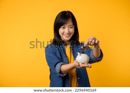 Happy asian young woman wearing yellow t-shirt denim shirt saving digital crypto currency to piggy bank isolated on yellow background. Digital money saving investment concept.