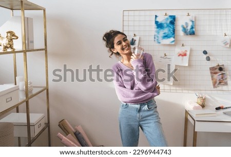 Delighted young woman in purple cardigan and jeans with closed eyes standing in light room near wall with inspiration board with various pictures while holding plastic cup