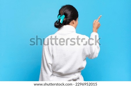 young hispanic woman standing and pointing to object on copy space, rear view. bathrobe concept