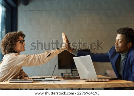 Successful young male and female colleagues giving each other high-five over workplace with laptops, nobepads and paper documents