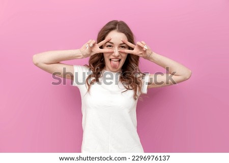 young cute woman shows tongue and makes peace gesture with hands on pink isolated background, girl holds fingers near eyes and poses