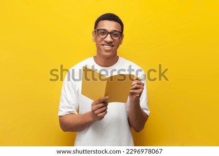guy african american student in white t-shirt and glasses writes in notebook and reads book on yellow isolated background, smart man holds book Royalty-Free Stock Photo #2296978067