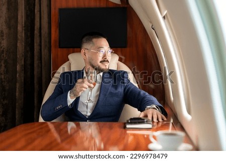 successful Asian businessman in suit and glasses sits in private jet and drinks champagne, Korean manager in business clothes flies in business class and looks out the window, luxury lifestyle Royalty-Free Stock Photo #2296978479