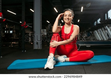 athletic woman in red sportswear sits on yoga matte in black gym and smiles, girl does yoga and warms up, portrait of attractive woman in fitness club Royalty-Free Stock Photo #2296984849