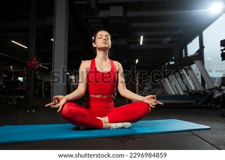 athletic woman in red sportswear sits on yoga matte in black gym and meditates, girl does yoga, attractive woman in fitness club in lotus position Royalty-Free Stock Photo #2296984859