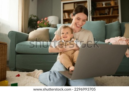 Cute baby boy and his mother sitting on the floor in the living room and using laptop