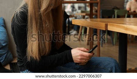 Unrecognizable young woman in coffee shop waiting for order with phone on knees. Faceless female with long brown hair sits at empty table type on smartphone, cars and people in street in background.