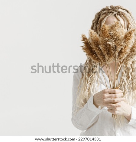 Woman holding pampas grass in front of face, light background, no face trend concept. Female with long curly hair dreadlocks hiding behind bouquet dry wild flower. Creative autumn visual. Fall concept