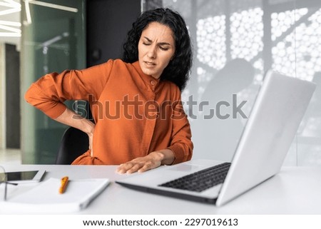 Overtired hispanic woman working late, business woman having severe back pain, female worker inside office sitting at table using laptop at work Royalty-Free Stock Photo #2297019613