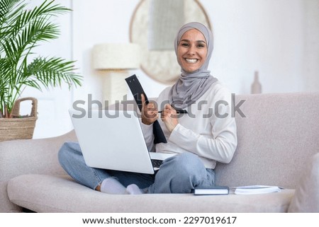 Young female student of oriental origin in hijab studying from home. Sitting on the sofa in the lotus position with a notebook and writing with a pen in a notebook, folder. Smiling at the camera.