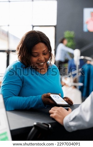 African american shopper buying fashionable clothes paying with smartwatch at fashion store counter desk, mobile phone with nfc and credit card. Woman shopping for formal wear in modern boutique