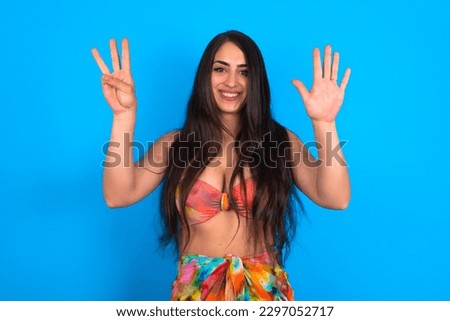 beautiful brunette woman wearing swimwear over blue background showing and pointing up with fingers number eight while smiling confident and happy.