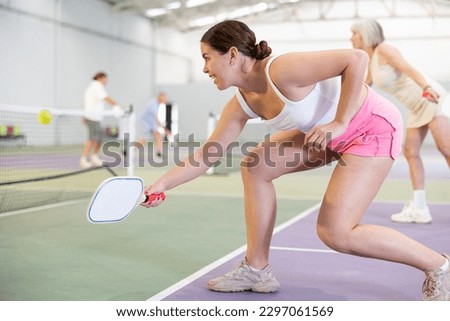 Concentrated fit young girl pickleball player waiting to receive serve, ready to strike and return ball to opponent field on indoor court.. Royalty-Free Stock Photo #2297061569