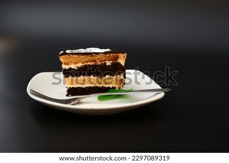 A piece of biscuit layer cake with chocolate and mint leaves on a white ceramic plate and a fork on a black background. Close-up.