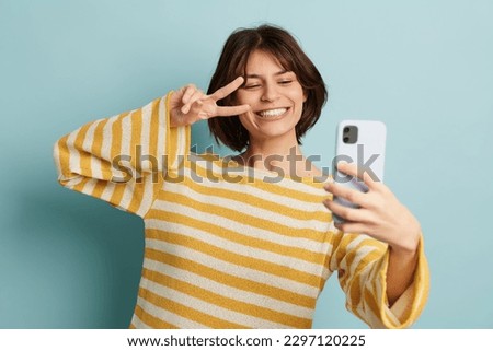 Optimistic young female in striped sweater smiling and showing sign while taking selfie against blue background