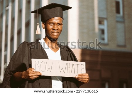 Portrait of disgruntled African guy standing with cardboard poster on street near building looking for job. University or college graduating student in gown and cap. Employment issue concept.  Royalty-Free Stock Photo #2297138615