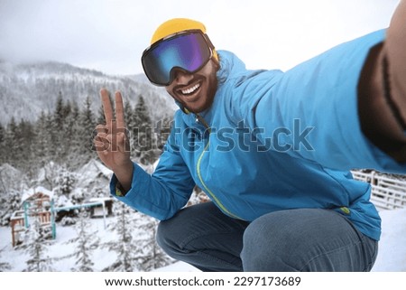 Smiling young man in ski goggles taking selfie and showing peace sign in mountains