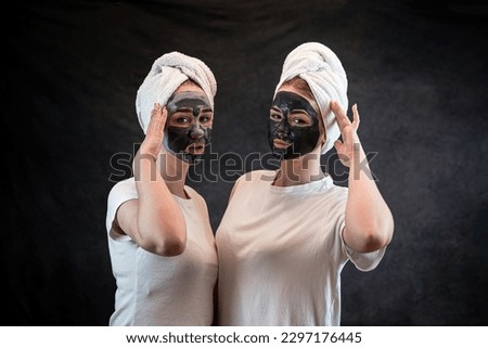 Girls friends in white towel applying black mud clay facial mask isolated on black. Anti age treatment, skincare