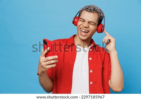 Young cheerful happy fun vivid man of African American ethnicity 20s he wearing red shirt headphones listen music sing song use mobile cell phone isolated on plain pastel light blue cyan background Royalty-Free Stock Photo #2297185967