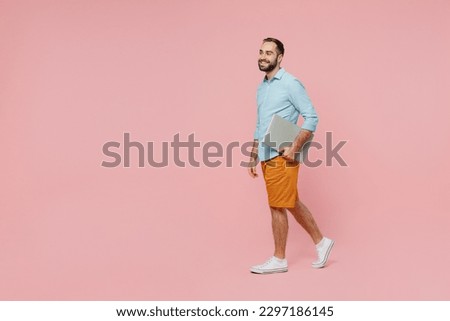 Full body side view young smiling man 20s wear classic blue shirt hold closed laptop pc computer walking going isolated on plain pastel light pink background studio portrait. People lifestyle concept