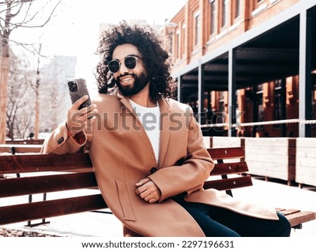 Handsome smiling model.  Arabian man dressed in brown coat clothes. Fashion male in the street. Sitting on a bench. Holding smartphone, looks at cellphone screen, use phone apps. Talks at video call