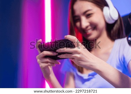 Winning video game on cellphone. Young asian pretty woman sitting on chair holding smartphone in living room. Happy female Pro Streamer chinese wearing headphone playing game online with neon light.