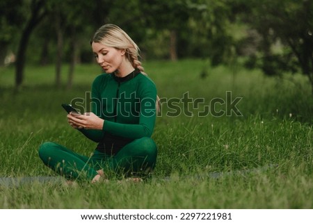 Yoga in the park with sunlight. A young woman in a lotus position sits on green grass holding a phone in her hand. The concept of digital transformation.