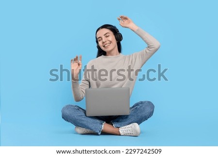 Happy woman with laptop listening to music in headphones on light blue background